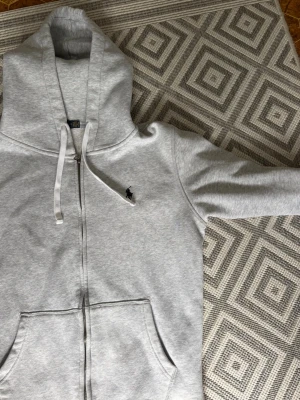 Grå zip hoodie från Polo Ralph Lauren - Snygg ljusgrå hoodie med dragkedja från Polo Ralph Lauren. Klassisk design med huva och vita dragsnören, samt broderad logga på bröstet. Två stora fickor framtill. Perfekt för en chill och stilren look.