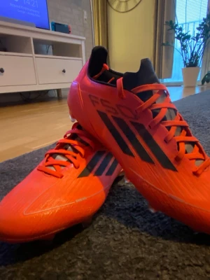 Röda Adidas F50 fotbollsskor - Säljer ett par röda Adidas F50 fotbollsskor med svarta och silvriga detaljer samt klassiska tre ränder på sidan. Skorna har snörning, låg profil och är tillverkade i syntetmaterial för lätt vikt och bra bollkontroll. Perfekta för dig som vill ha speed på planen.
