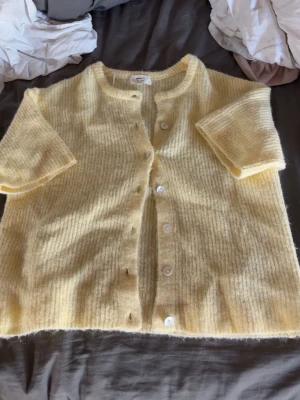 Gul fluffig kofta med kort ärm - Mysig ljusgul kofta med korta ärmar och rund hals. Koftan är stickad i ett fluffigt material och har knappar framtill. Perfekt för lager på lager och ger en söt, avslappnad look.