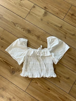Vit croppad blus med volang - Supersöt vit croppad blus med breda korta ärmar och volangdetaljer. Toppen har smockad midja, liten rosett framtill och är perfekt för sommarens alla tillfällen!