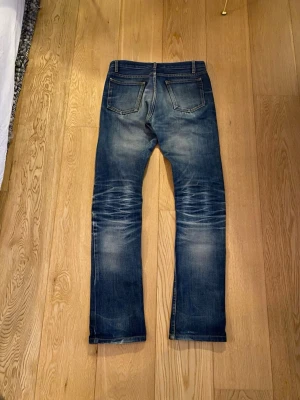 Blå slitna jeans från Nudie Jeans - Säljer ett par blå jeans från Nudie Jeans med snyggt slitna detaljer och klassisk femficksmodell. Jeansen har raka ben och tydliga whiskers vid knäna och baksidan. Tillverkade i denim med en cool tvättad look.
