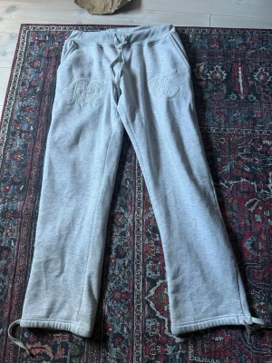 Grå mjukisbyxor med brodyr - Säljer ett par ljusgrå mjukisbyxor med broderade bokstäver på framsidan. Byxorna har dragsko i midjan och raka ben med justerbara snören vid bensluten. Perfekta för chill dagar och riktigt sköna att ha på sig.