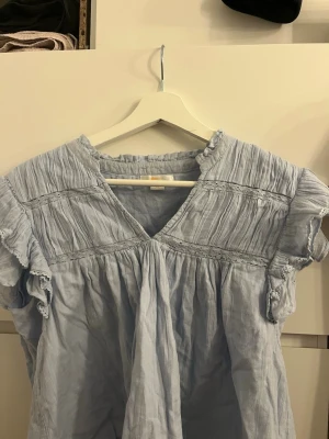 Ljusblå blus med spetsdetaljer - Söt ljusblå blus i tunn, luftig bomull med v-ringning och spetsdetaljer över bröstet och axlarna. Blusen har korta, volangprydda ärmar och ett lätt rynkat tyg som ger en avslappnad känsla. Perfekt till jeans eller kjol.