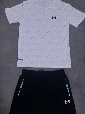 Under armour vit/svart tränings sett   -  Snyggt träningsset från Under Armour med en vit t-shirt med diskret mönster och svart logga på bröstet. Tillhörande svarta shorts med dragkedjefickor och vit logga. Perfekt för gymmet eller bara en varm sommar Settet är påväg kontakta för intresse