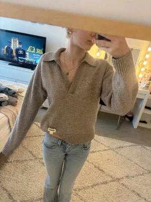 Beige stickad tröja med krage - Säljer en beige stickad tröja med bred krage och djup v-ringning. Tröjan har långa ärmar, ribbade muddar och en relaxed fit. Perfekt för lager på lager och enkel att styla till jeans eller kjol.