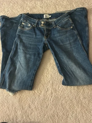 Blå raka jeans från 157 - Säljer ett par klassiska blå jeans från 157 med raka ben och femficksmodell. Jeansen har dubbla knappar i midjan, ljus tvätt och slitningar vid bensluten för en avslappnad look. Perfekta till en casual outfit.
