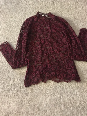 Vinröd spetsblus med hög krage - Vinröd blus i genombruten spets med blommigt mönster och hög krage. Blusen har långa ärmar och en feminin, elegant look. Perfekt för dig som vill sticka ut med en unik och trendig stil.