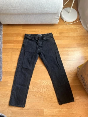 Svarta jeans från Jack & Jones - Säljer ett par svarta raka jeans från Jack & Jones. Klassisk femficksmodell med knappgylf och normal passform. Jeansen är tillverkade i bomull och har en stilren look som passar till det mesta.