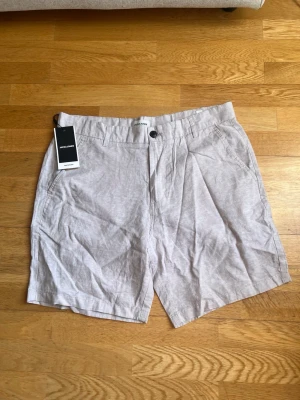 Beige linneshorts från Jack & Jones - Snygga beige shorts från Jack & Jones i en luftig linnemix. Klassisk passform med fickor fram och bak, bälteshällor och knappstängning. Perfekta för varma dagar när du vill ha en clean och avslappnad stil.