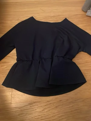 Svart peplum topp med lång ärm - Snygg svart peplum topp med lång ärm och markerad midja. Toppen har en rund halsringning och volang nedtill som ger en feminin siluett. Perfekt att styla med jeans eller kjol för en trendig look.
