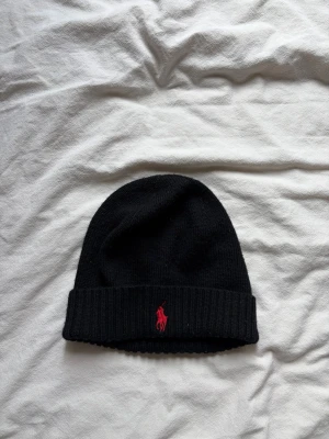 Svart mössa från Polo Ralph Lauren - Snygg svart mössa från Polo Ralph Lauren i mjuk merinoull. Klassisk ribbad kant med röd broderad logga framtill. Perfekt accessoar för kalla dagar och enkel att matcha med olika stilar.