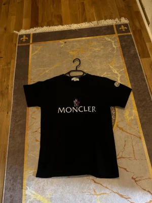 Svart Moncler t-shirt med logga - Snygg svart t-shirt från Moncler med stor vit logga och röd detalj på bröstet. Klassisk passform och korta ärmar. Moncler-märke även på ärmen. Perfekt för dig som gillar exklusiva streetwear-plagg.