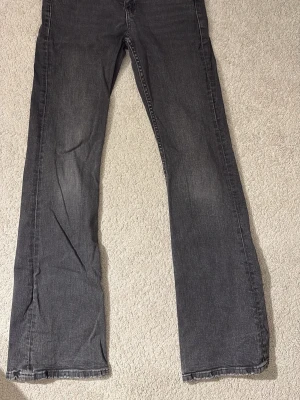 Grå bootcut jeans med ficklock - Säljer ett par grå jeans med bootcut-modell och snygga slitningar. Jeansen har ett ficklock med knappdetalj bak och är tillverkade i klassisk denim. Perfekta för dig som gillar en avslappnad men trendig stil. De är lowwaist och bootcut! De passar även folk som är längre! De passade mig som var 155 ungefär. De är i bra skick! Det enda som är lite slitet är fickorna (se bild 2) och längst ner på jeansen (se bild 4) men det syns ej när man har på sig dem! Skriv till mig för frågor!