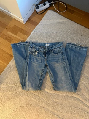 Ljusblå bootcut jeans med råa kanter - Säljer ett par ljusblå bootcut jeans med råa, fransiga benslut och klassisk femficksdesign. Jeansen har en avslappnad passform och är perfekta för en trendig och avslappnad look. Materialet är denim i bomull.