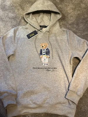 Grå hoodie Polo Bear Ralph Lauren - Snygg grå hoodie från Polo Ralph Lauren med den ikoniska Polo Bear-trycket framtill. Klassisk modell med huva och dragsko samt ribbade muddar vid ärmar och midja. Tillverkad i mjukt bomullsmaterial för maximal komfort. Perfekt för en avslappnad och trendig look. (Rep)