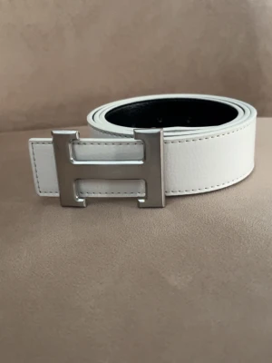 Vitt H’e’r’m’e”s skärp med H spänne - Snyggt vitt skärp från H’e’r’m’e”s med H spänne. Bältet har en slät yta och svarta insidan. Perfekt accessoar för att lyfta vilken outfit som helst.