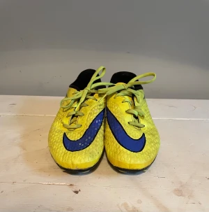 Nike Hypervenom gula fotbollsskor - Säljer ett par Nike Hypervenom fotbollsskor i en riktigt schysst gul färg med blåa detaljer och svart insida. Skorna har klassisk snörning och dobbar för spel på gräs. Ovandelen är tillverkad i syntetmaterial med struktur för bättre bollkontroll.
