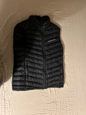Svart dunväst från Peak Performance - Snygg svart dunväst från Peak Performance med diskret logga på bröstet. Västen har dragkedja framtill, ståkrage och två sidofickor med dragkedja. Quiltad design med horisontella sömmar och lätt vaddering för extra värme.