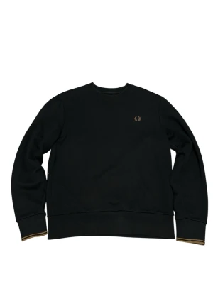 Svart sweatshirt från Fred Perry - Klassisk svart sweatshirt från Fred Perry med rund halsringning och diskret guldfärgad logga broderad på bröstet. Tröjan har ribbade muddar med gulddetaljer vid ärmslut och nederkant. Perfekt för en clean och stilren look.