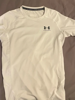 Vit träningströja från Under Armour - Säljer en vit, tight och kortärmad träningströja från Under Armour med svart logga på bröstet. Tillverkad i ett tekniskt funktionsmaterial som andas, perfekt för intensiva träningspass eller löpning.