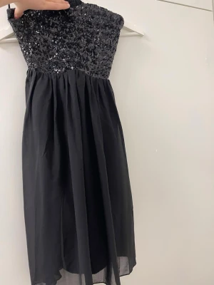 Svart axelbandslös paljettklänning - Säljer en svart axelbandslös klänning med glittrande paljetter över bysten och ett lätt, flödande tyg i kjolen. Klänningen har smock i ryggen för skön passform och är perfekt för dig som vill sticka ut. Materialet känns lätt och festligt.