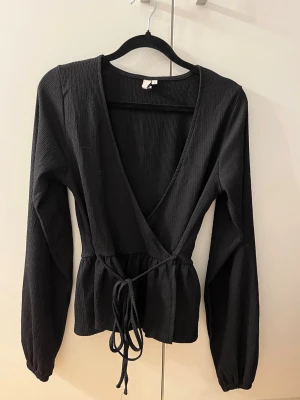 Svart omlottopp från Nelly - Svart omlottopp från Nelly med djup v-ringning (passar bra med ett vitt linne under!!💕) och långa ärmar. Toppen har knyt i midjan och är tillverkad i ett ribbat material som ger en snygg struktur. 