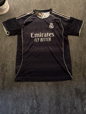Real Madrid   - Snygg mörkblå Real Madrid matchtröja från Adidas med klubbmärke på bröstet och 'Emirates Fly Better' tryckt framtill. Tröjan har korta ärmar, ljusa detaljer längs sidor och axlar samt klassiska Adidas-ränder. Perfekt för fotbollsträning eller support.