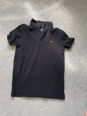 Svart piké från Lyle & Scott - Klassisk svart pikétröja från Lyle & Scott med korta ärmar och gul logga på bröstet. Tröjan har knappar vid halsen och är gjord i mjuk bomull. Perfekt till jeans eller shorts för en clean och enkel stil.