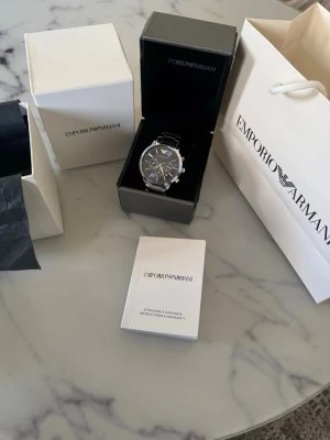 Emporio Armani silver/blå klocka - Stilren klocka från Emporio Armani med silverfärgat boett och svart läderarmband. Klassisk rund urtavla med tydliga markeringar och diskreta detaljer. Perfekt accessoar för dig som gillar en elegant och tidlös look.