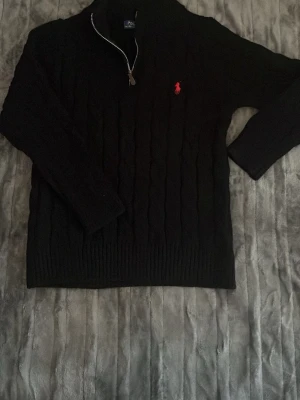 Svart stickad tröja Polo Ralph Lauren - Svart stickad tröja från Polo Ralph Lauren med halv dragkedja och röd broderad logga på bröstet. Tröjan har kabelstickat mönster, ribbade muddar och hög krage. Perfekt för en stilren och klassisk look.