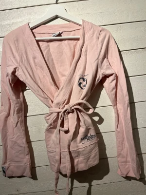 Ljusrosa omlott tröja  Better Bodies - Mysig ljusrosa omlott tröja  från Better Bodies med långa ärmar och knytskärp i midjan. Tryck på bröstet och fickan med logga och texten 'Wellness'. Tillverkad i mjukt bomullsmaterial, perfekt för chilliga morgnar eller påväg till yoga passet . Strl M 