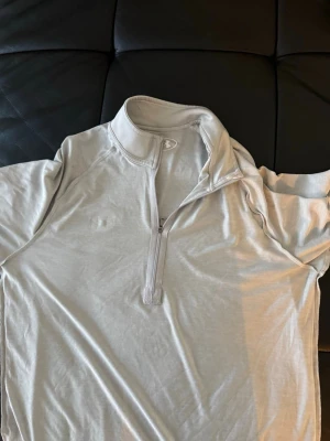 Träningströja Under Armour - Säljer en ljusgrå långärmad träningströja från Under Armour med halv dragkedja framtill och hög krage. Tillverkad i ett mjukt och lätt funktionsmaterial som andas bra, perfekt för träning eller löpning.