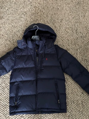 Mörkblå dunjacka Polo Ralph Lauren - Snygg mörkblå dunjacka från Polo Ralph Lauren med huva och dragkedja. Jackan har två framfickor med dragkedja och en liten röd logga på bröstet. Perfekt för kalla vinterdagar och riktigt stilren design.