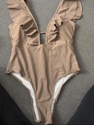 Beige baddräkt med volangdetaljer - Snygg beige baddräkt med djup V-ringning och volangdetaljer längs axelbanden. Tillverkad i 85% polyester och 15% elastan för en stretchig och bekväm passform. Perfekt för strand eller poolhäng med trendig look.