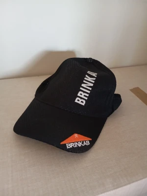 Svart keps från BRINKAB - Snygg svart keps från BRINKAB med vit broderad text på framsidan och en orange detalj med logga på skärmen. Klassisk böjd skärm och justerbar baksida. Perfekt för dig som vill ha en stilren och enkel look.