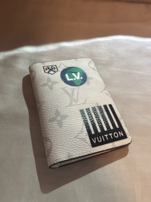 Lv plånbok - Säljer en vit plånbok från Louis Vuitton med monogrammönster och coola tryck i svart, grönt och blått. Plånboken har ett modernt grafiskt mönster med texten 'VUITTON' och 'L.V.' samt detaljer i olika färger. Tillverkad i tåligt lädermaterial.