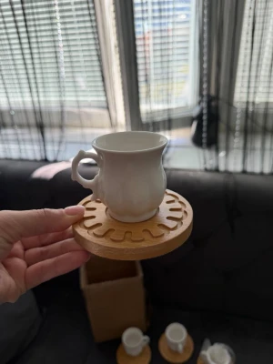 Set med vita koppar och träunderlägg - Säljer ett set med fem vita koppar i keramik med snyggt formade handtag och tillhörande runda underlägg i ljust trä med dekorativt mönster. Perfekt för fika eller te. Kopparna har en klassisk design och underläggen ger en naturlig touch till dukningen.