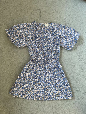 Blommig blå kortklänning neo noir - Supersöt kortklänning från neo noir med blått och vitt blommönster. Klänningen har puffiga korta ärmar, rund halsringning och smock i midjan för en snygg siluett. Perfekt för vår och sommar, tillverkad i mjuk bomull.