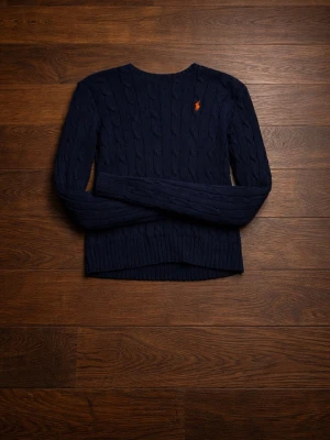 Mörkblå kabelstickad tröja Polo Ralph Lauren - Kabelstickad mörkblå tröja från Polo Ralph Lauren med rund halsringning och lång ärm. Klassisk orange logga broderad på bröstet. Tröjan har ribbade muddar vid ärmslut och nederkant, perfekt för lager på lager och en stilren look.