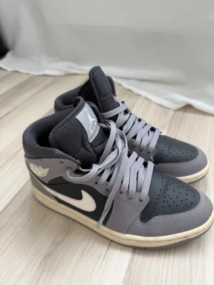 Air Jordan 1 Mid grå/lila och svart - Snygga Air Jordan 1 Mid sneakers i grått och svart med vita detaljer och klassisk vinge-logga på sidan. Skorna har en vit sula och är gjorda i läder med textildetaljer. Perfekta för dig som gillar streetwear och vill ha en ikonisk look.