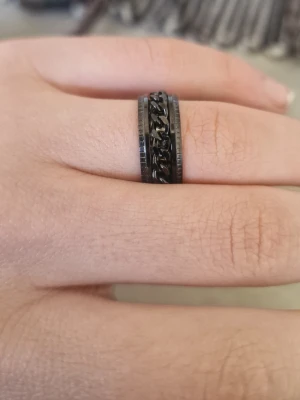 Svart ring med kedjedetalj - Snygg svart ring i metall med en cool kedjedetalj runt hela ringen. Ringen har en bred design och en modern, industriell look som sticker ut. Perfekt för dig som gillar accessoarer med edge och vill ha något unikt på fingret.