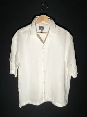 Vit linneskjorta från H&M - Säljer en stilren vit skjorta från H&M i luftigt linne. Skjortan har korta ärmar, klassisk krage och knappar framtill. Perfekt för varma dagar och enkel att matcha med olika outfits.