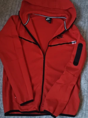 Röd Nike hoodie med dragkedja - Snygg röd hoodie från Nike med svart dragkedja och svarta detaljer. Hoodien har en klassisk huva, svarta kantband och en liten ficka med dragkedja på ärmen. Nike-logga broderad på bröstet. Perfekt för en sportig och avslappnad stil. Vet inte om den är äkta.