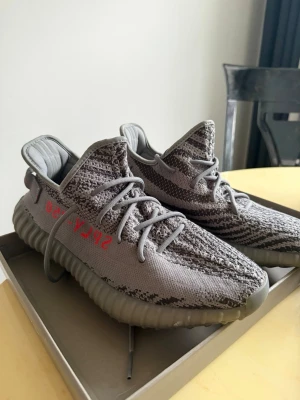 Yeezy Boost 350 V2 Beluga - Säljer ett par Yeezy Boost 350 V2 Beluga med grå och mörkgrå Primeknit-överdel, orange SPLY-350 text på sidan och genomskinlig grå sula med Boost-dämpning. Skorna har en ikonisk siluett och snörning framtill. Perfekta för dig som gillar streetwear och sneakers.