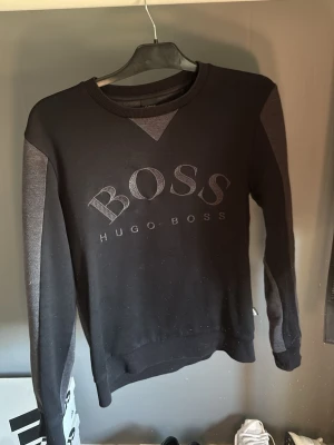 Svart och grå sweatshirt från Hugo Boss - Stilren svart och grå sweatshirt från Hugo Boss med stort BOSS-tryck på bröstet. Tröjan har rund halsringning, ribbade muddar och kontrastfärgade ärmar. Perfekt för en avslappnad och trendig look.