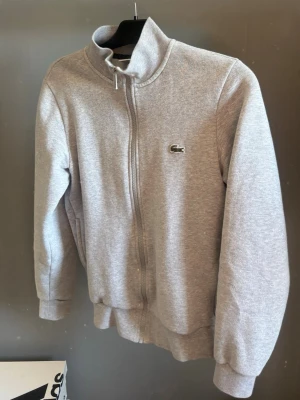 Grå ziptröja från Lacoste - Snygg grå ziptröja från Lacoste med klassisk ståkrage och dragkedja hela vägen. Tröjan har det ikoniska krokodilmärket på bröstet och ribbade muddar vid ärmar och nederkant. Perfekt för en chill och sportig look.