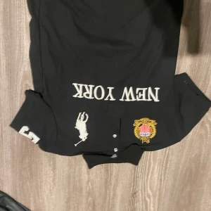 Svart pikétröja Polo Ralph Lauren M - Svart pikétröja från Polo Ralph Lauren med broderad logga, New York-tryck och guldfärgad patch på bröstet. Klassisk krage, knappar framtill och siffra på ärmen. Perfekt för dig som gillar sportig och stilren look. Den må vara av ursprung som skilljer sig från raph lauren men ja vet inte