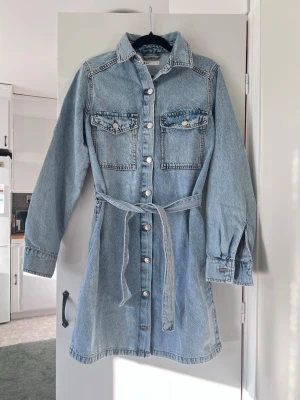 Ljusblå jeansklänning, storlek S - Säljer denna fina blåa jeansklänning med knappar och knytband från Gina Tricot. Blev aldrig använd, endast testad. Mjuk och härlig i materialet. Säljs ej längre.   Längd: 88cm