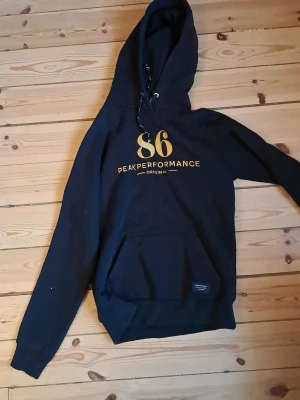 Svart hoodie från Peak Performance - Snygg svart hoodie från Peak Performance med gul text '86' och logga på bröstet. Klassisk känguruficka framtill, justerbar huva med snören och ribbade muddar. Perfekt för en avslappnad streetwear-look.