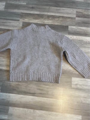 Grå ribbstickad tröja med hög krage - Mysig grå stickad tröja med ribbstickat mönster och hög krage. Tröjan har långa ärmar och en något boxig, croppad passform. Perfekt för lager på lager och enkel att matcha med jeans eller kjol.